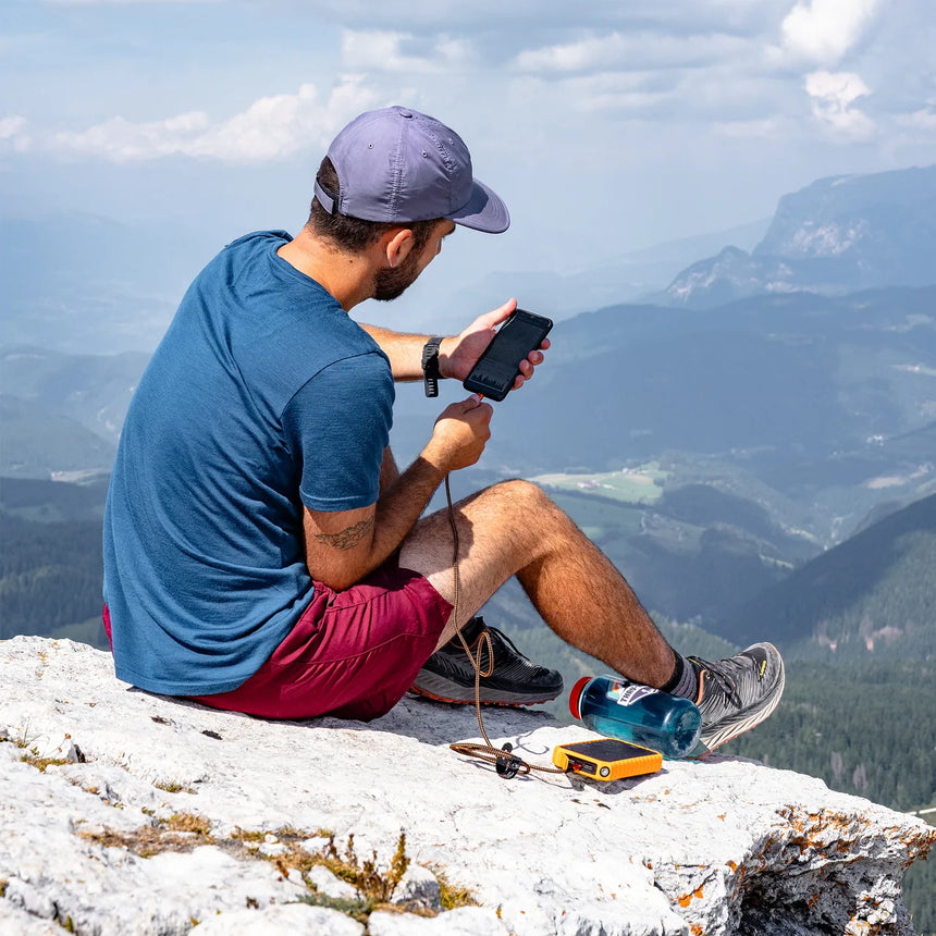 Man sitting on a mountain top using a smartphone with a scenic view of mountains in the background.